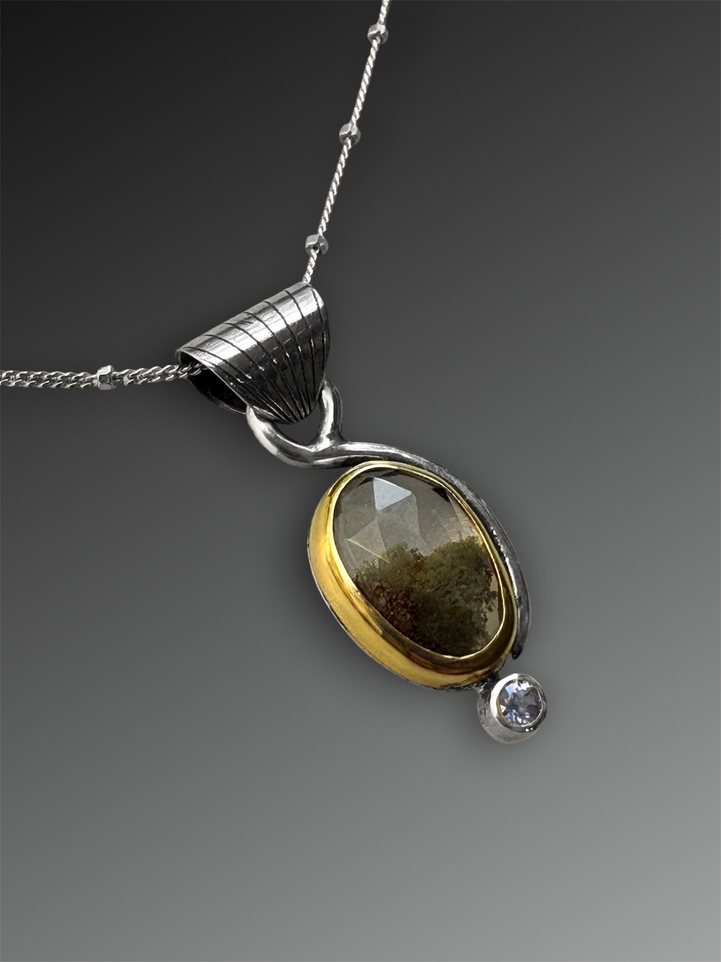 Silver and gold two-stone pendant with a stone above that looks like it has moss inside it, and a small tanzanite below, on a gray background