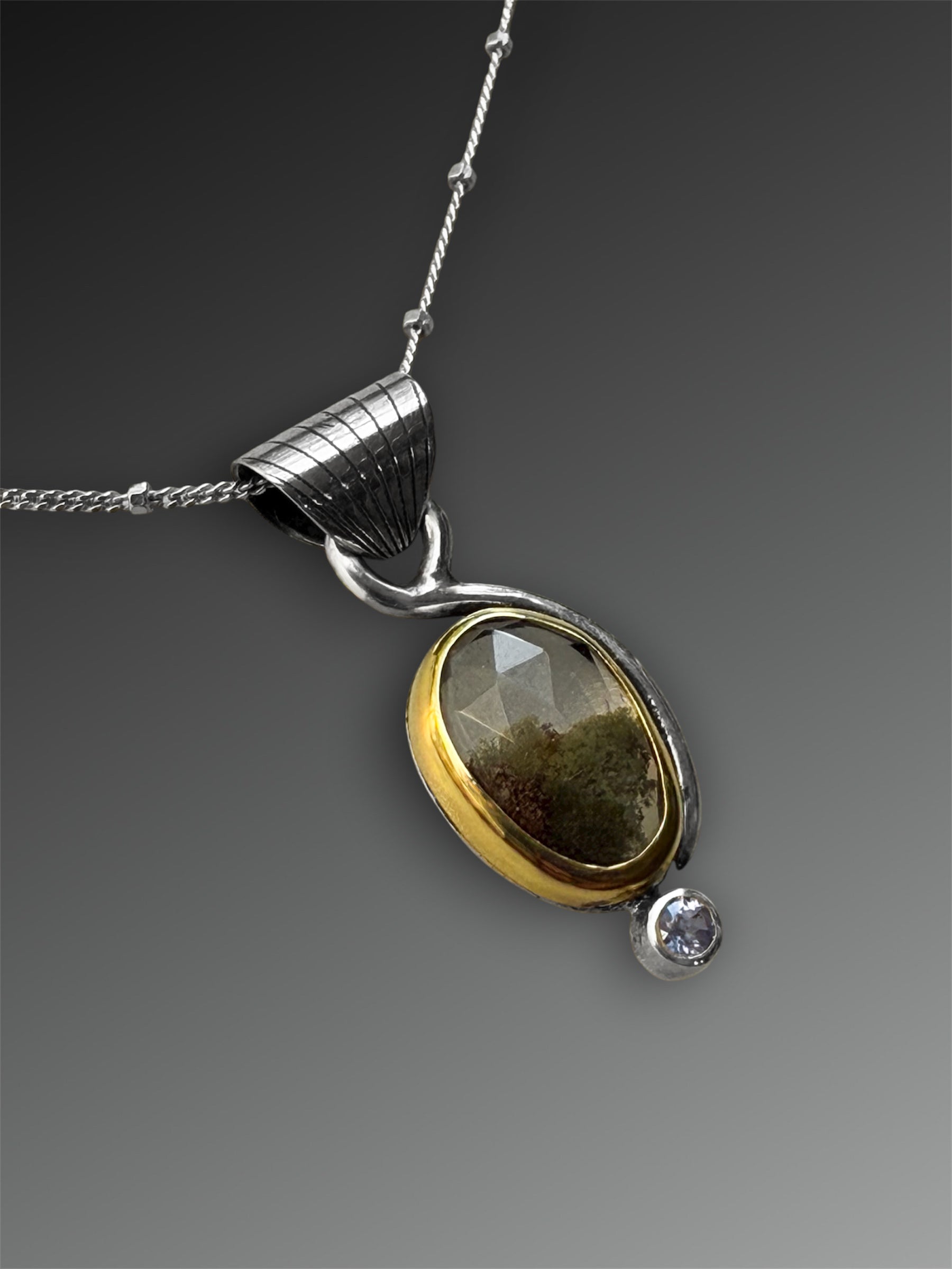 Silver and gold two-stone pendant with a stone above that looks like it has moss inside it, and a small tanzanite below, on a gray background